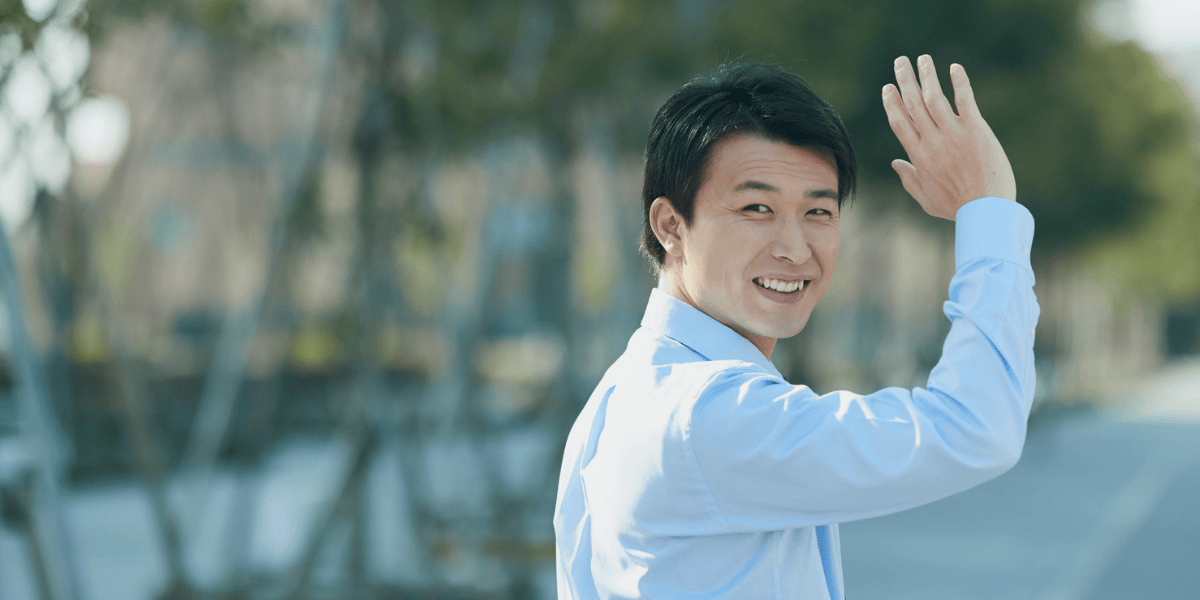 Junger Mann im hellblauen Hemd winkt lächelnd auf einer Straße mit Bäumen im Hintergrund