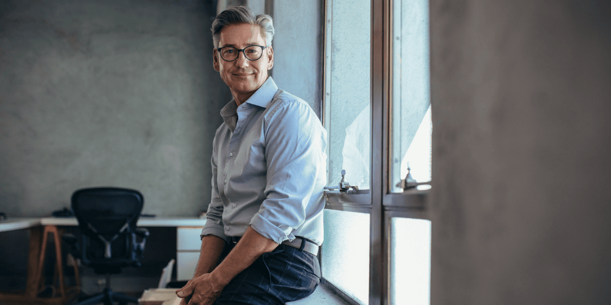 Älterer Mann mit Brille und grauem Hemd sitzt auf einer Fensterbank im Büro, Schreibtisch und Bürostuhl im Hintergrund