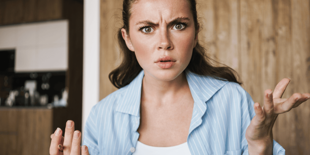 Porträt einer Person mit zurückgebundenem braunem Haar und blau gestreiftem Hemd, Stirn gerunzelt, Hände angehoben, Innenraum mit Holzwand im Hintergrund.