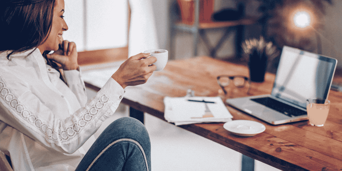 Frau mit weißer Bluse sitzt am Schreibtisch, hält eine Tasse Kaffee und blickt auf einen Laptop; auf dem Holztisch liegen Notizbuch, Stift, Brille und Untertasse.