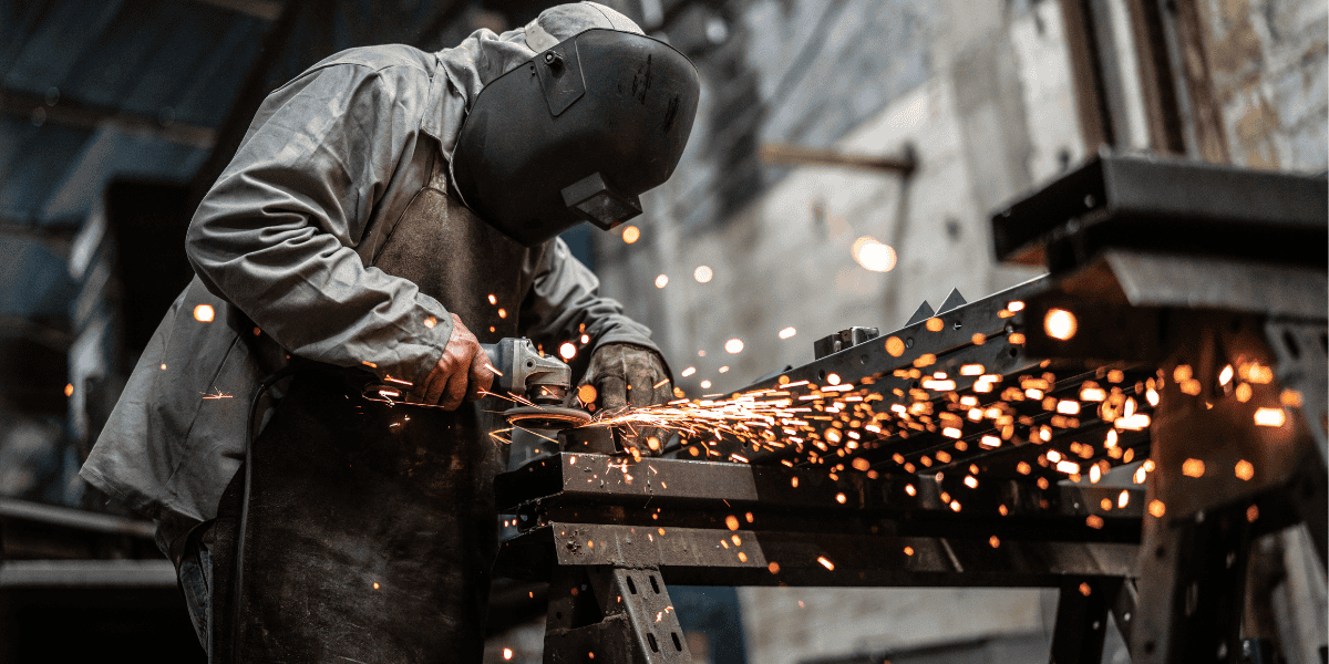 Schutzbekleidete Person mit Schweißhelm schleift Metall an Werkbank; Funken sprühen in Industrieumgebung.