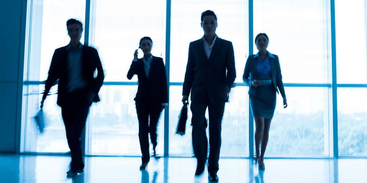 Vier Geschäftsleute gehen als Silhouetten vor großen Fenstern in modernem Bürogebäude