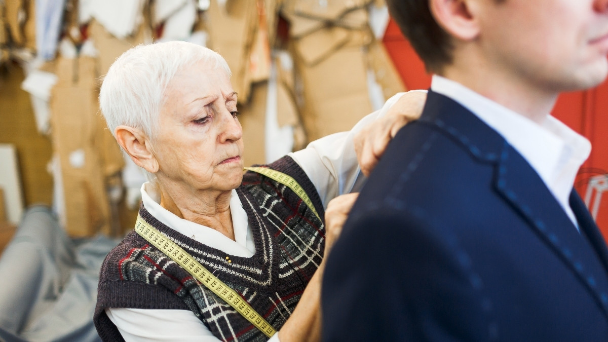 Ältere Schneiderin misst die Schulter eines Mannes im Anzug mit einem Maßband