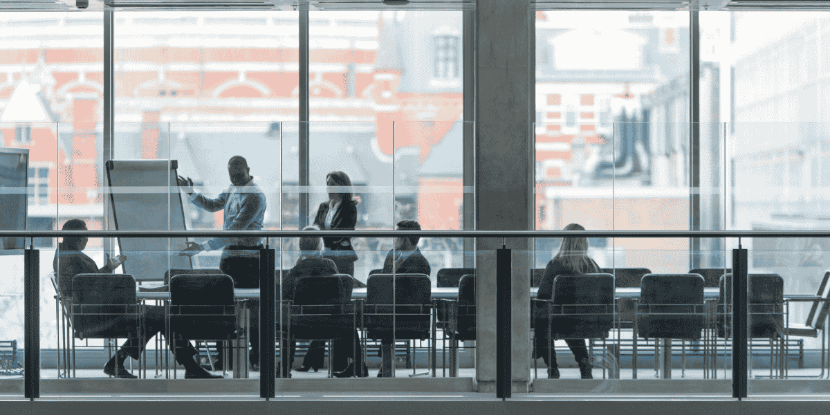 Besprechung in modernem Konferenzraum hinter Glaswand, Person präsentiert am Flipchart, mehrere Teilnehmende sitzen am Tisch, große Fenster mit Stadtansicht im Hintergrund.