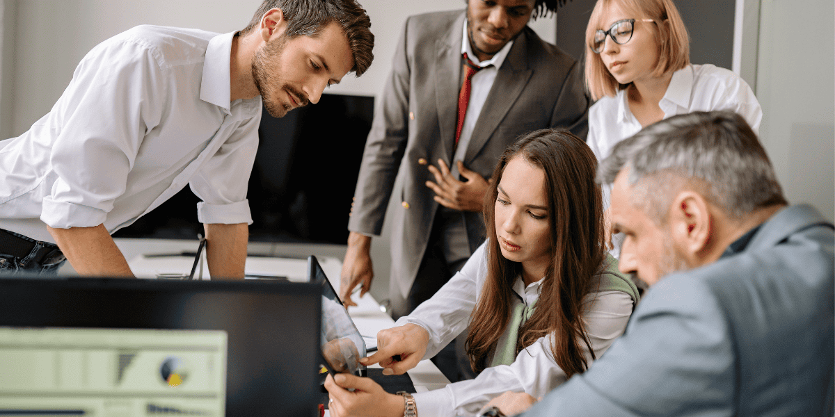 Fünf Personen im Büro beugen sich über Laptop und Dokumente und besprechen Inhalte gemeinsam mit neugieriger Mimik.