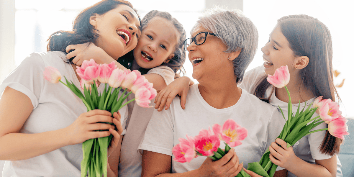 Vier Personen verschiedener Generationen sitzen eng zusammen, lachen und halten rosa Tulpensträuße in den Händen, helles Wohnraum-Ambiente im Hintergrund.