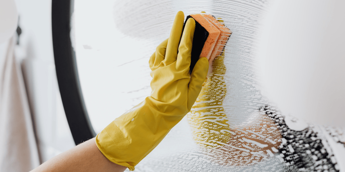 Hand mit gelbem Gummihandschuh reinigt eine Glasfläche mit Schwamm und Seifenwasser, Spiegel und Badezimmerumgebung im Hintergrund.