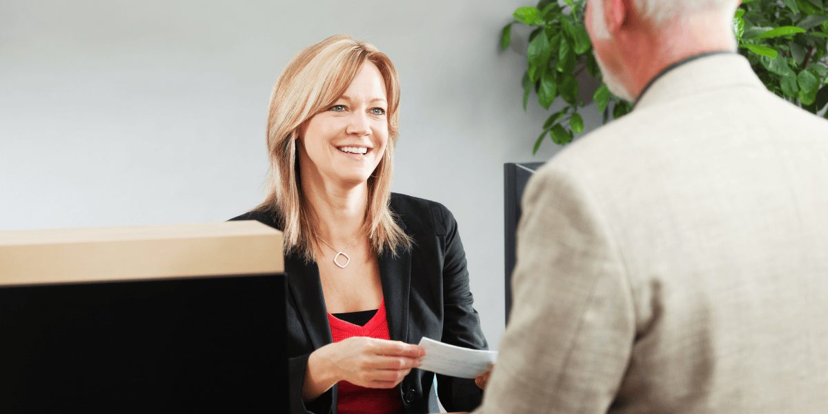 Frau im schwarzen Blazer überreicht einem Mann im Büro eine Karte am Schreibtisch, Beratungsgespräch am Empfangstresen.