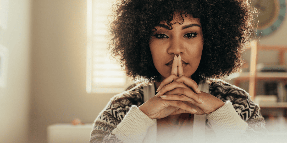 Porträt einer Person mit lockigem Haar, Hände vor dem Gesicht gefaltet, Zeigefinger an den Lippen, in Innenraum mit Fenster und Regal im Hintergrund.