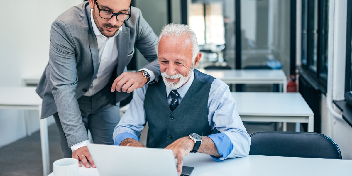 Zwei Personen im Büro arbeiten gemeinsam an einem Laptop, eine sitzend mit Weste und Krawatte, die andere im Anzug danebenstehend; Tasse auf dem Tisch.
