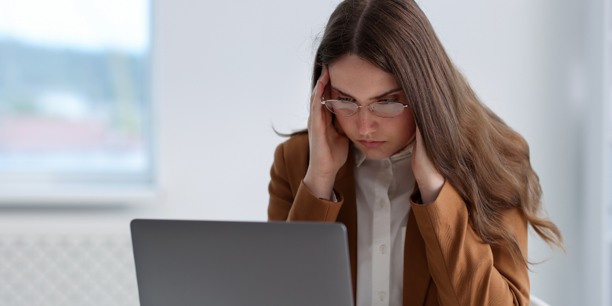 Person mit Brille und braunem Blazer sitzt vor einem Laptop und hält die Hände an die Schläfen in einem hellen Büro.