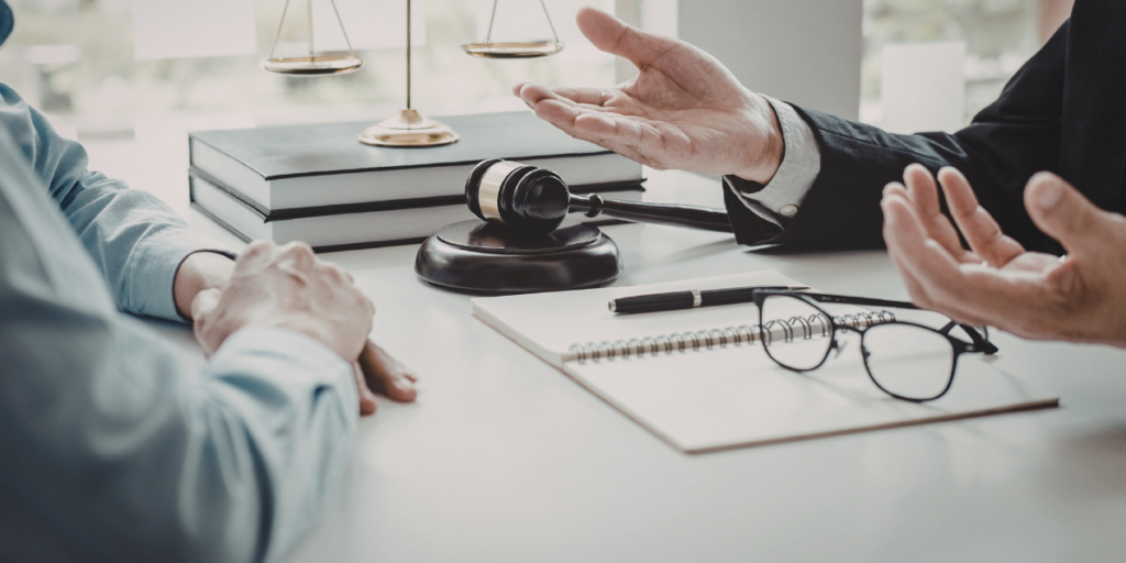 Beratungsgespräch am Schreibtisch mit Richterhammer, Gesetzbüchern, Waage der Justiz, Notizblock und Brille – zwei Personen diskutieren eine Angelegenheit.