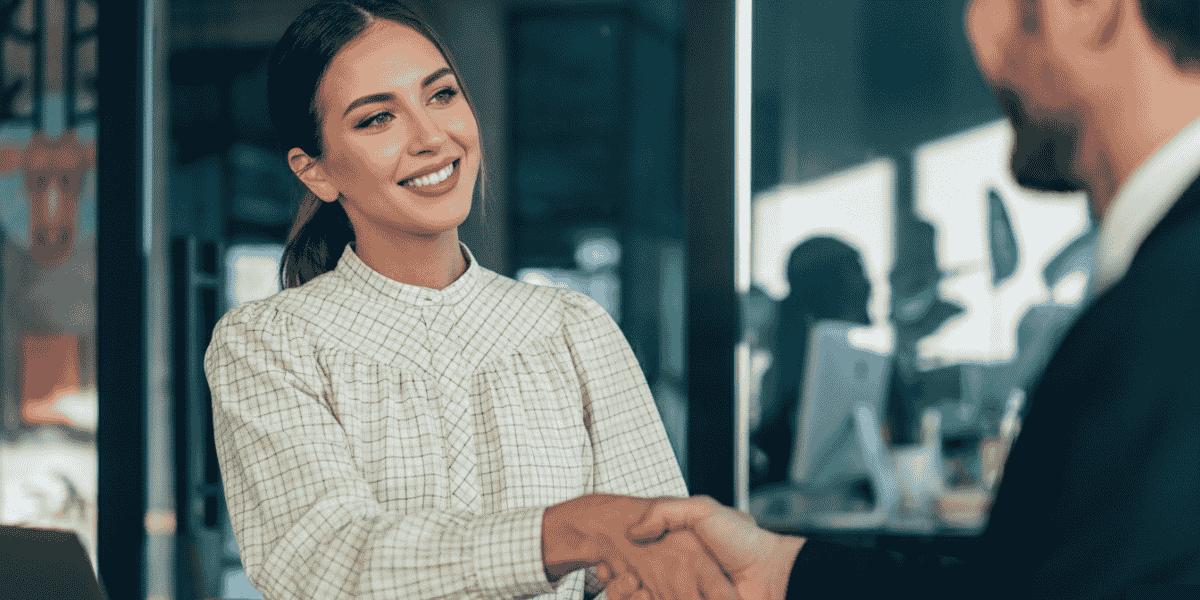 Geschäftsfrau im Büro schüttelt einem Mann im Anzug die Hand, Glaswand und Computerarbeitsplatz im Hintergrund