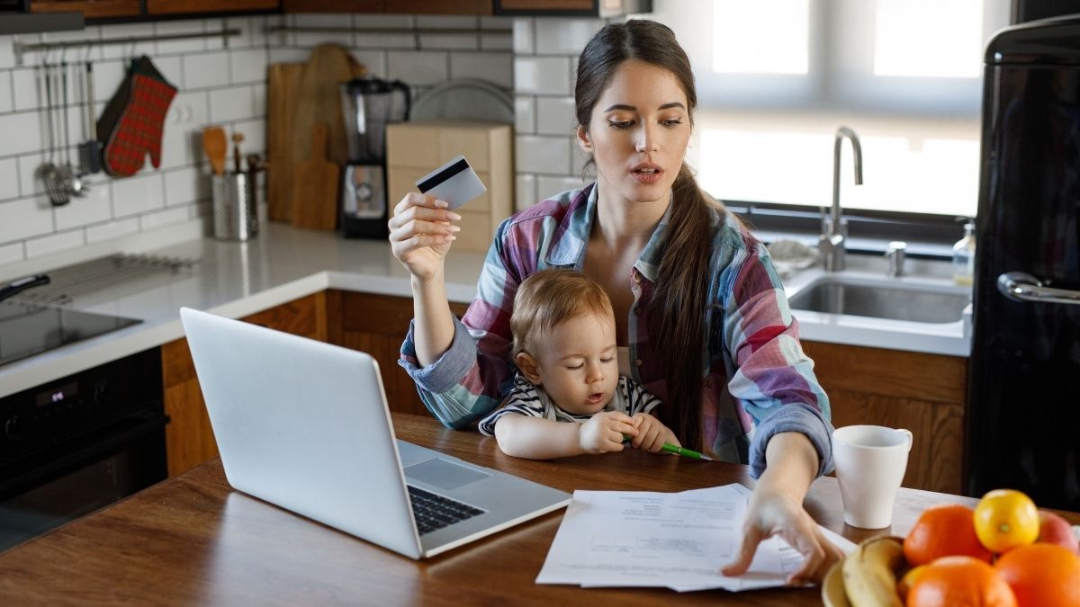 Junge Mutter sitzt mit Kleinkind am Küchentisch, hält eine Kreditkarte in der Hand