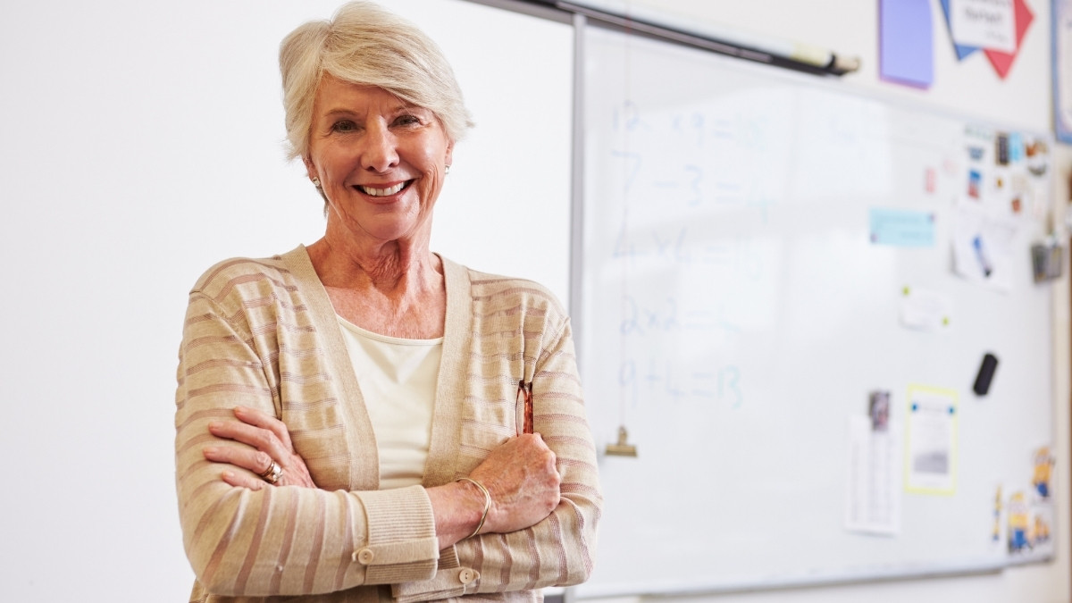 Ältere Frau steht vor einer Tafel im Klassenzimmer und lächelt mit verschränkten Armen