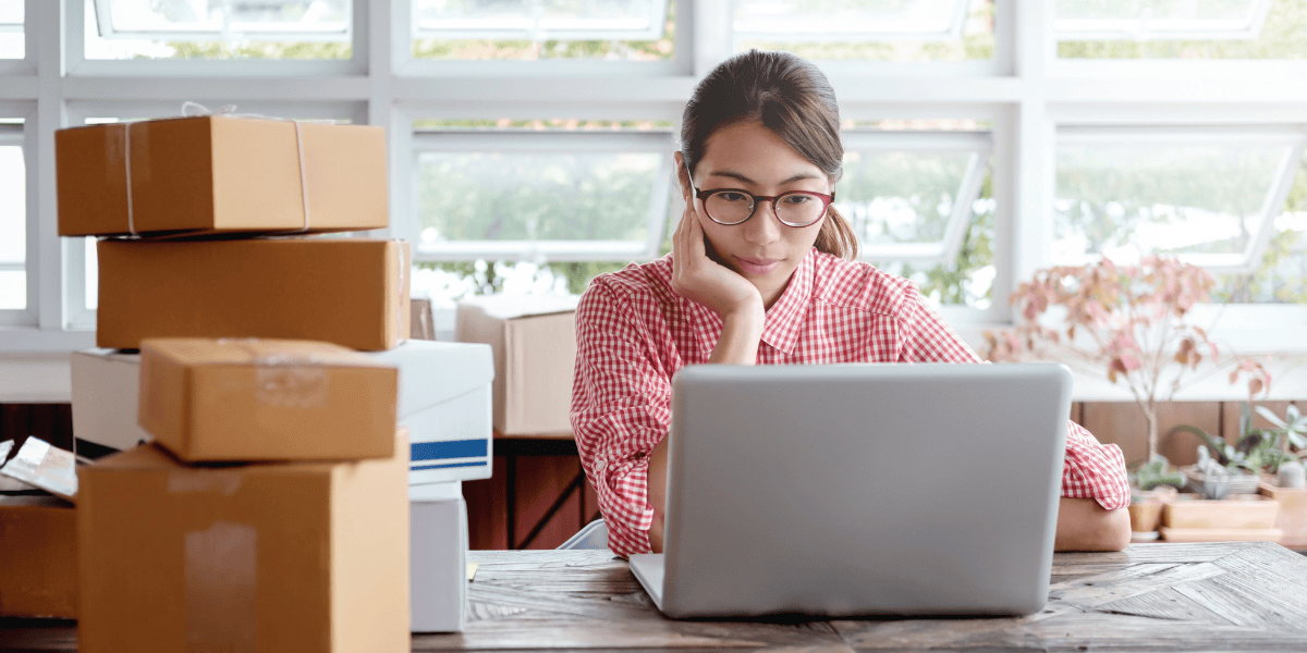 Frau mit Brille im karierten Hemd arbeitet am Laptop neben gestapelten Versandkartons in hellem Büro mit Fenstern, ein Online-Shop oder Versandarbeitsplatz.