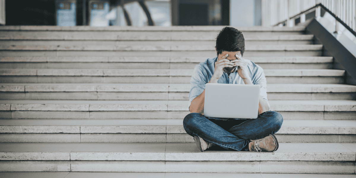 Person sitzt im Schneidersitz auf einer breiten Außentreppe aus Beton, Laptop auf dem Schoß, Hände vor dem Gesicht, trägt Jeans, Sneaker und helles Hemd.