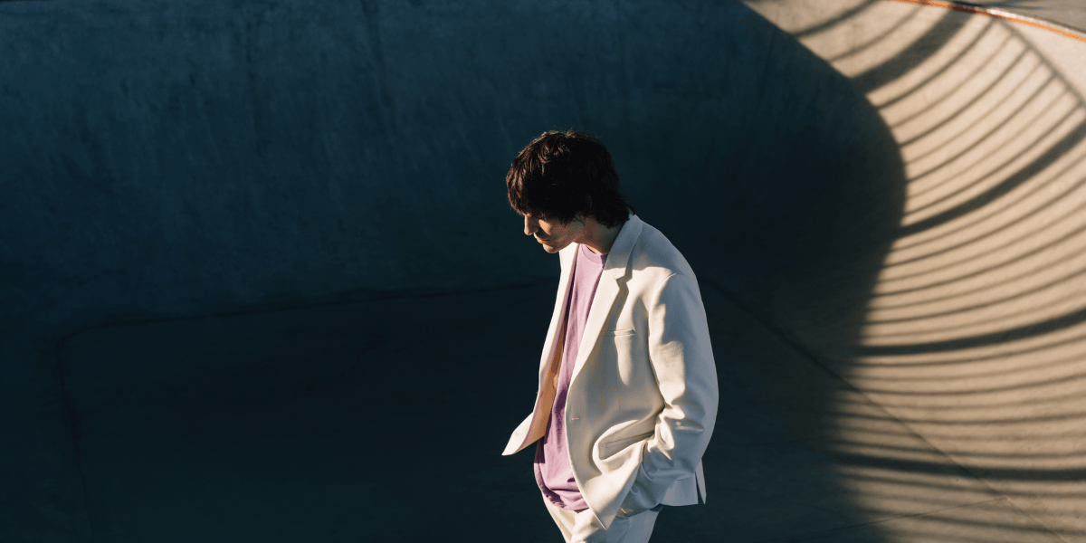 Person mit dunklem, mittellangem Haar im weißen Anzug und violettem Shirt steht in einer runden Betonarchitektur, starkes Sonnenlicht erzeugt lineare Schatten eines Geländers an der Wand.