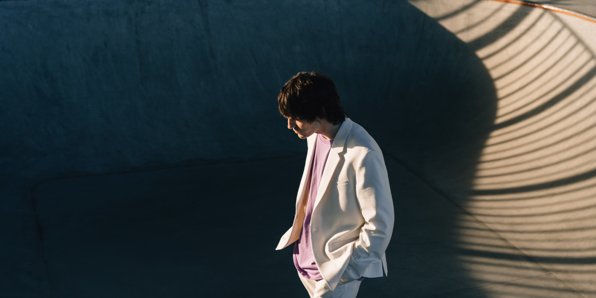 Person mit dunklem, mittellangem Haar im weißen Anzug und violettem Shirt steht in einer runden Betonarchitektur, starkes Sonnenlicht erzeugt lineare Schatten eines Geländers an der Wand.