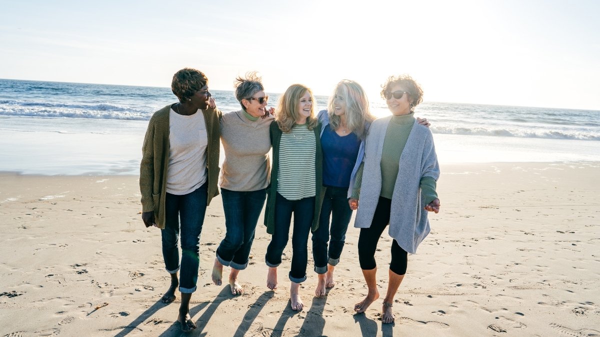Fünf Frauen gehen gemeinsam lachend am Strand entlang, Arm in Arm, mit dem Meer im Hintergrund