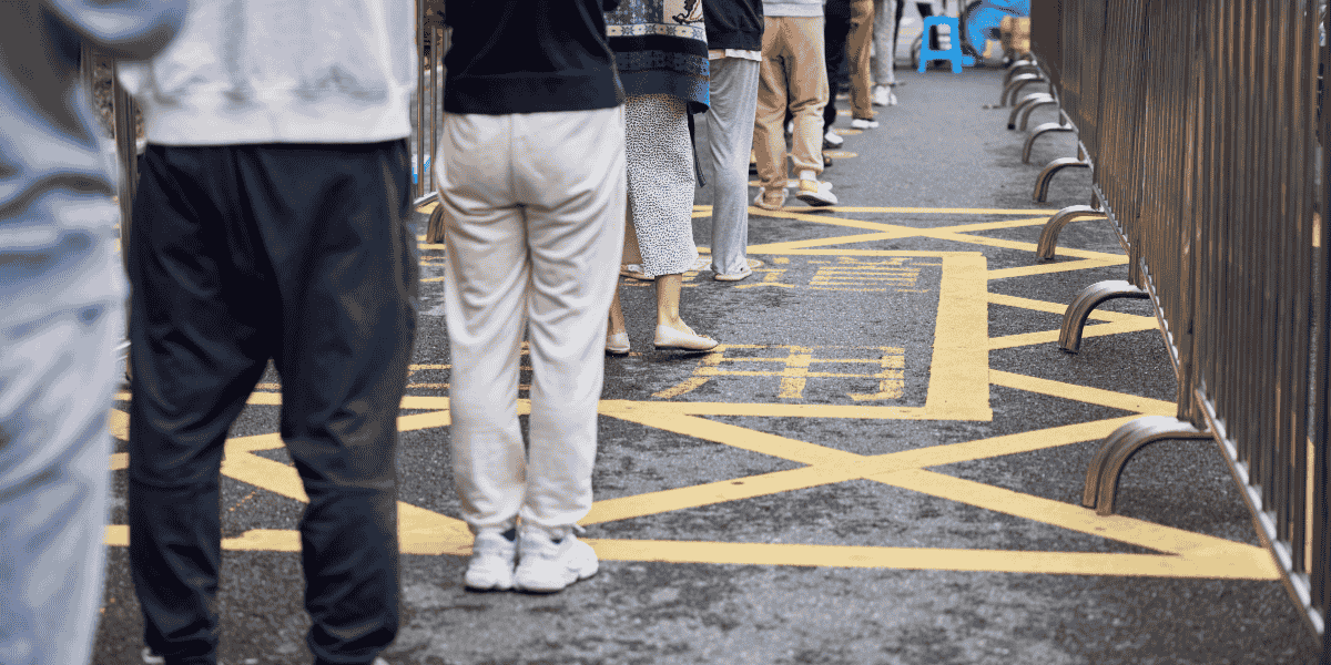 Warteschlange von Personen zwischen Metall-Absperrgittern auf Asphalt mit gelber Kreuzschraffur-Straßenmarkierung im Außenbereich.