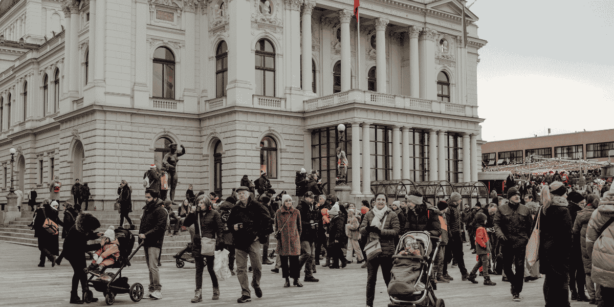 Historisches Gebäude mit Säulenfassade, große Menschenmenge auf dem Platz davor, Personen mit Kinderwagen und Winterkleidung, städtische Szene bei Tageslicht. Opernhaus Zürich.