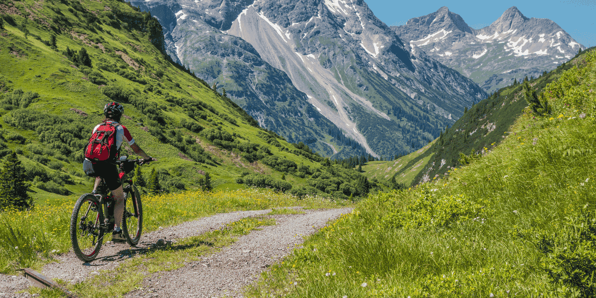 Radfahrende Person mit rotem Rucksack auf Schotterweg in grüner Berglandschaft, umgeben von alpinen Gipfeln unter blauem Himmel