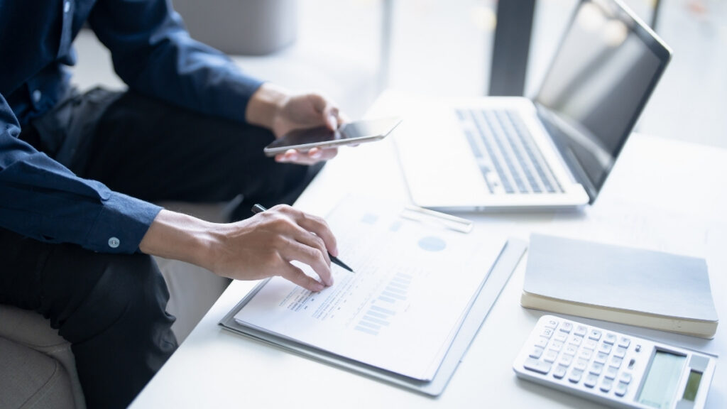 Person analysiert Dokumente mit Diagrammen am Schreibtisch und nutzt gleichzeitig ein Smartphone neben Laptop und Taschenrechner