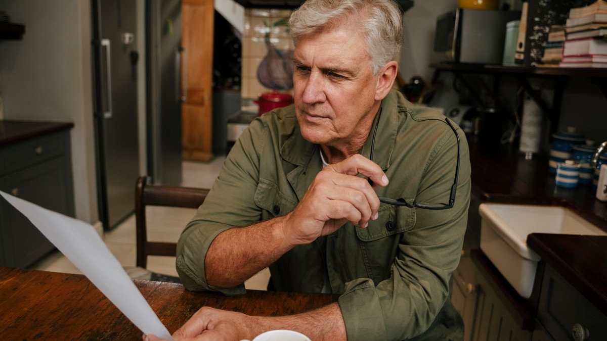 Älterer Mann sitzt am Tisch in der Küche und betrachtet nachdenklich ein Dokument in seiner Hand