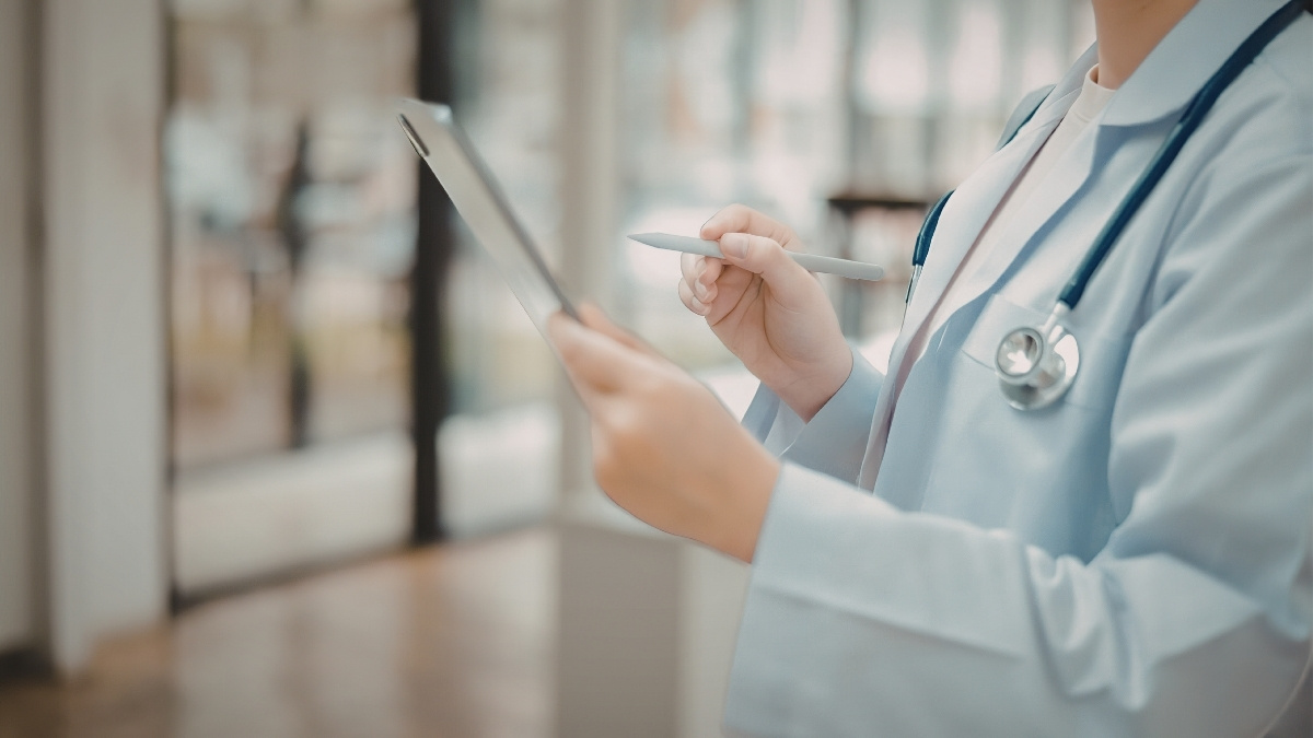 Medizinische Fachkraft in Kittel hält ein Tablet und einen Stift, mit Stethoskop um den Hals in einer hellen Umgebung