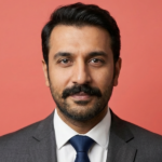 Porträt eines Erwachsenen mit dunklem Haar und Schnurrbart, Anzug mit Krawatte, rötlicher Hintergrund.