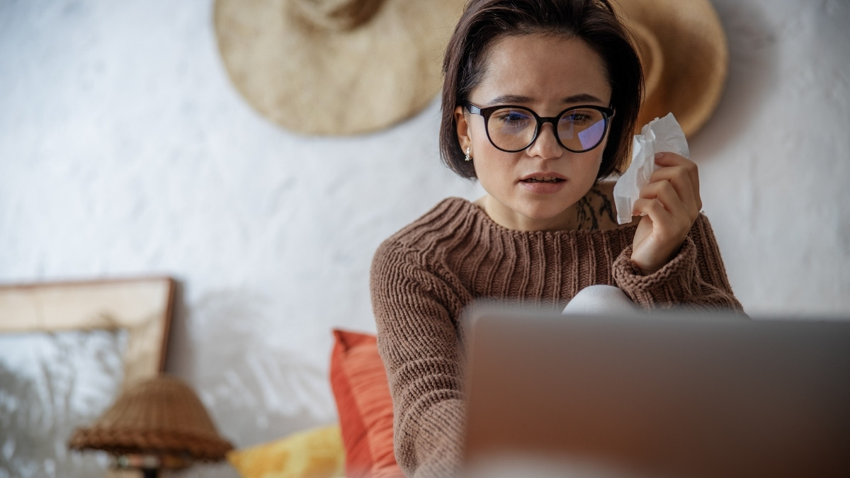 Frau mit Brille sitzt vor einem Laptop und hält ein Taschentuch, wirkt besorgt