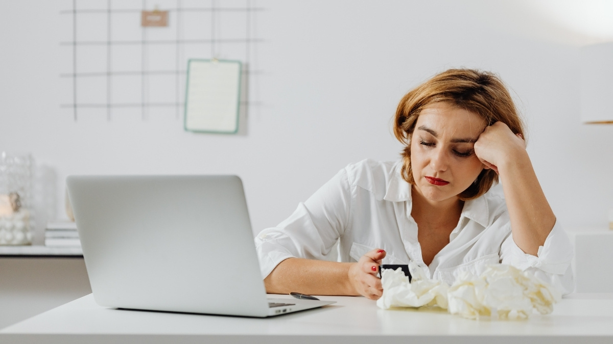 Erschöpfte Frau sitzt am Schreibtisch vor einem Laptop, umgeben von Taschentüchern, und blickt müde auf ihr Smartphone