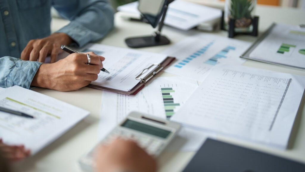 Person bearbeitet Finanzunterlagen mit Stift und Taschenrechner, umgeben von Diagrammen und Dokumenten auf dem Schreibtisch