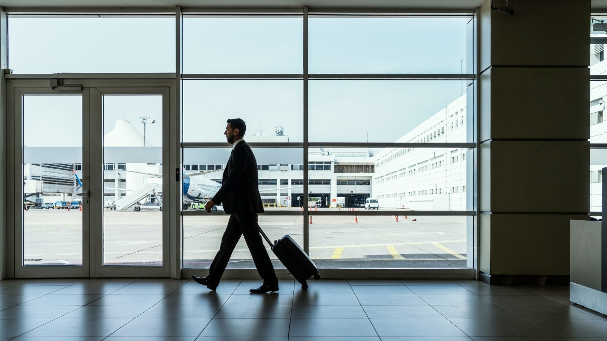 Geschäftsmann zieht einen Rollkoffer durch einen Flughafen und geht an großen Fenstern mit Blick auf das Rollfeld vorbei