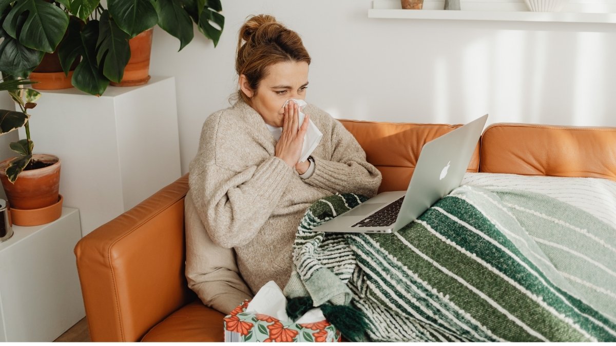 Erkältete Frau sitzt mit Decke auf dem Sofa, benutzt ein Taschentuch und arbeitet am Laptop, krank im Homeoffice