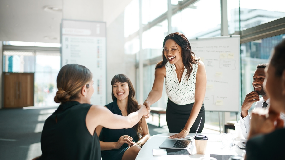Geschäftsfrauen begrüßen sich mit einem Handschlag in einem Meeting, während Kollegen lächelnd am Tisch sitzen