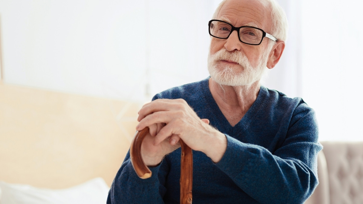Älterer Mann mit Brille sitzt zuhause und stützt sich nachdenklich auf einen Gehstock