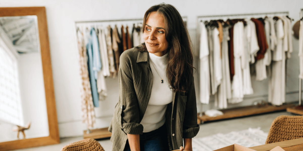 Frau steht in einem Geschäft zwischen Kleidung und schaut nachdenklich zur Seite.