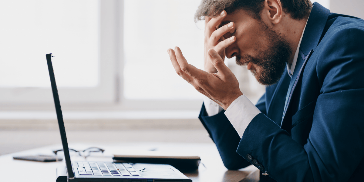 Person im Anzug sitzt am Schreibtisch vor Laptop, Hand an Stirn, Arbeitsumgebung mit Brille und Notizbuch