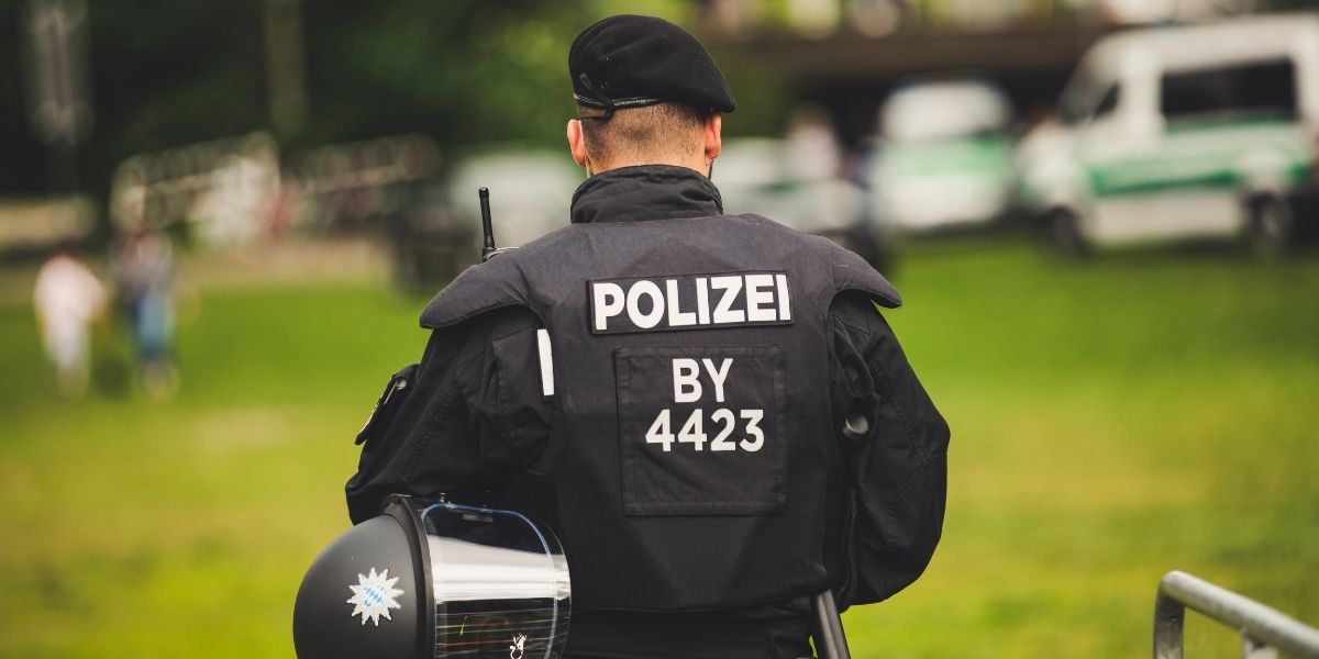Polizeibeamt*in in schwarzer Schutzkleidung von hinten, mit Helm und Aufschrift „POLIZEI BY 4423“, auf einer Grünfläche im Freien.