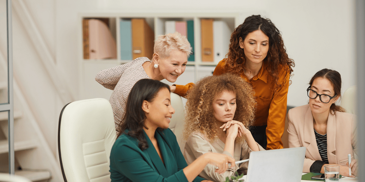 Fünf Frauen sitzen und stehen gemeinsam an einem Laptop im Büro und besprechen Inhalte.