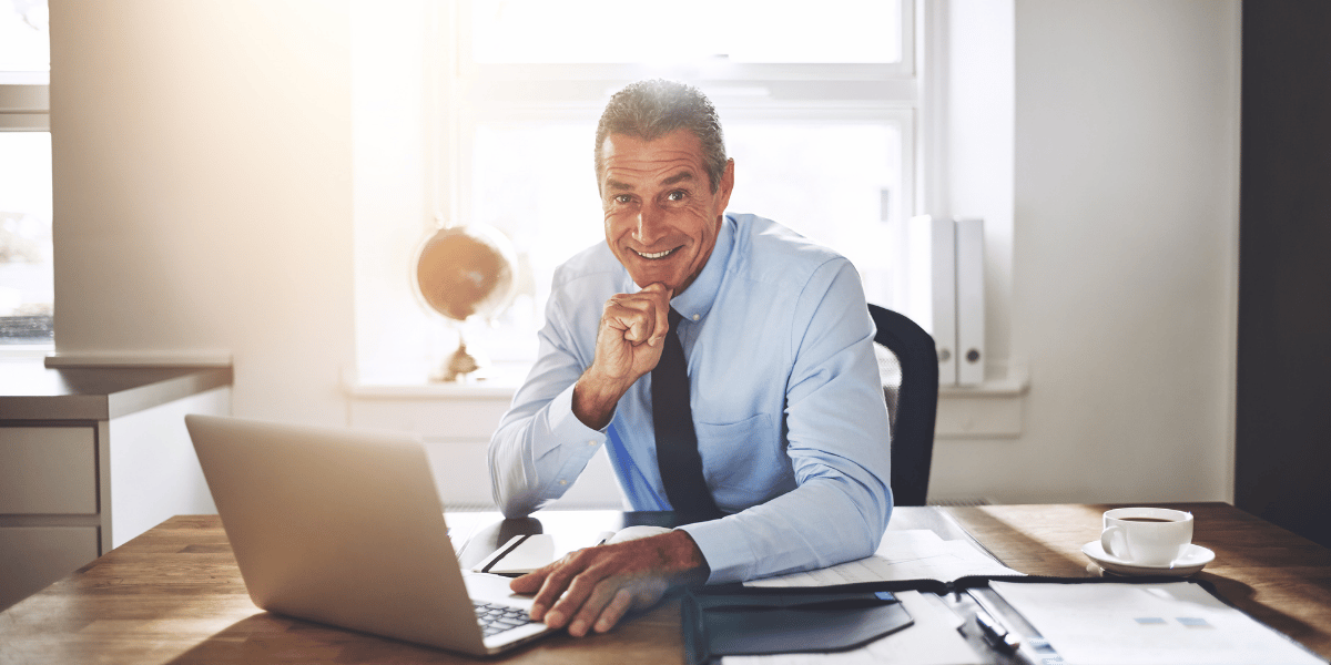 Person im Hemd und Krawatte sitzt am Schreibtisch vor Laptop, Hand am Kinn, Unterlagen und Kaffeetasse auf dem Tisch, helles Büro mit Fenster.