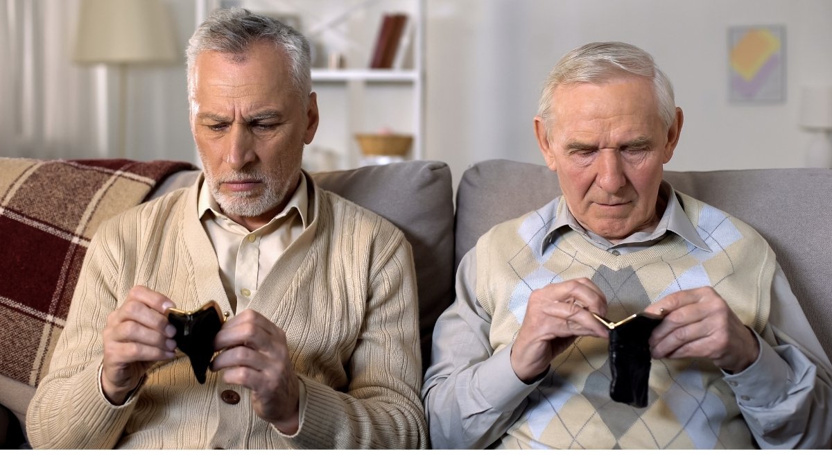 Zwei ältere Männer sitzen nebeneinander auf einem Sofa und schauen besorgt in ihre leeren Geldbörsen