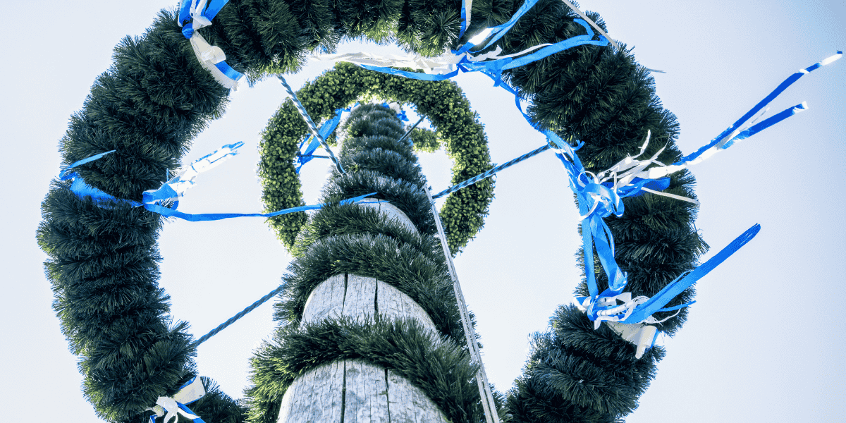 Alt-Text: Blick nach oben auf Holzmast mit grünen Girlandenringen und blau-weißen Bändern vor hellem Himmel.