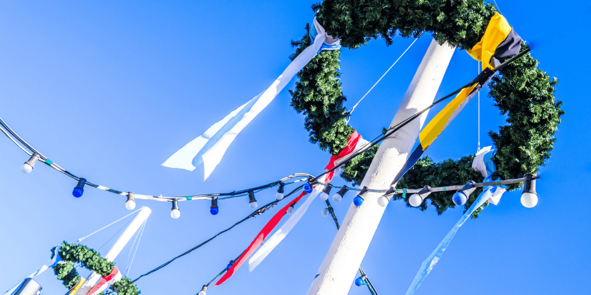 Blick nach oben auf weißen Mast mit grünem Kranz, bunten Bändern und Lichterkette vor blauem Himmel.