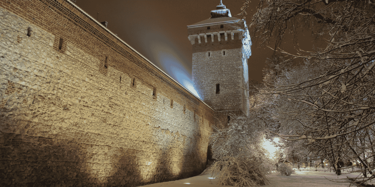 Beleuchtete mittelalterliche Steinmauer mit Turm bei Nacht, verschneite Bäume und Boden im Winter.
