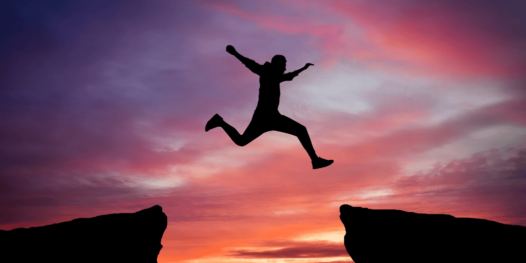 Silhouette einer Person springt zwischen zwei Felsen vor Sonnenuntergang.