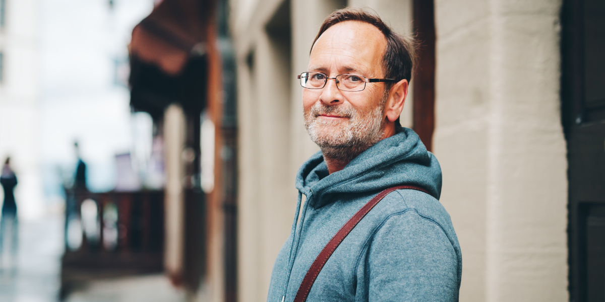 Mann mit Brille und grauem Hoodie steht seitlich in einer Straße, Schulterriemen sichtbar, unscharfer Hintergrund mit Gebäuden und Passanten.