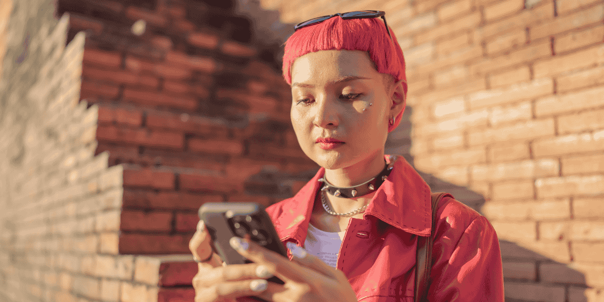 Person mit kurzen pinken Haaren und roter Jacke blickt auf ein Smartphone vor einer Ziegelwand im Sonnenlicht